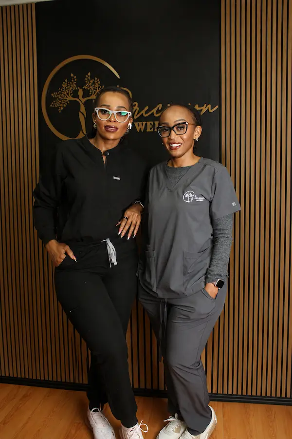 Two Precision Wellness team members in medical scrubs standing together in front of the clinic’s branded reception wall.